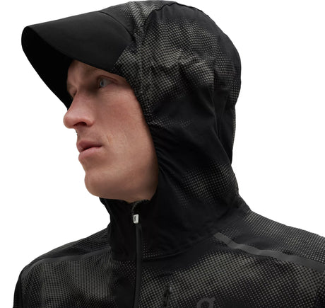 A man wearing a black reflective weather jacket with hoodie covering his head, zipper, and a logo on the chest.